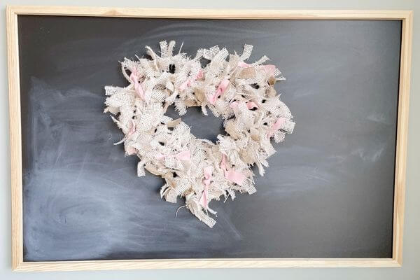 White burlap heart wreath with pink and brown ribbons hanging on chalkboard.