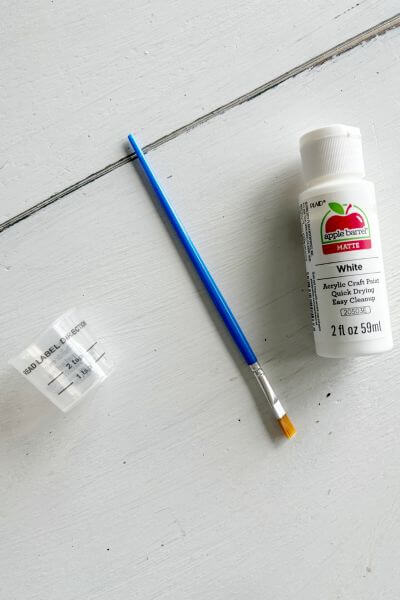 Supplies including a paintbrush, small measuring cup, and white acrylic paint. 