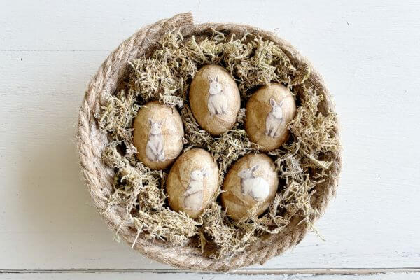 Five decoupaged eggs with vintage rabbit cut-outs sitting in a jute rope nest.