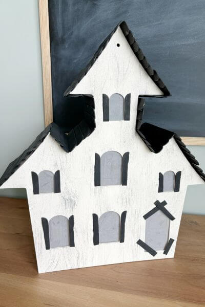 Wood haunted house with a roof, shutters and popsicle stick pieces glued around the door. 