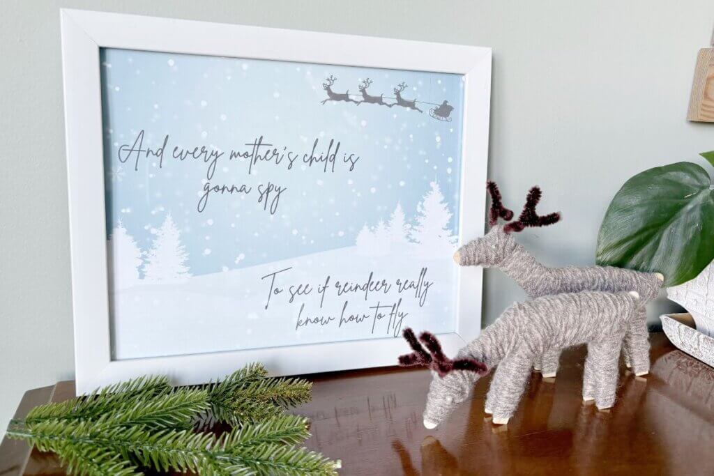 Two yarn reindeer next to a picture and some greenery.