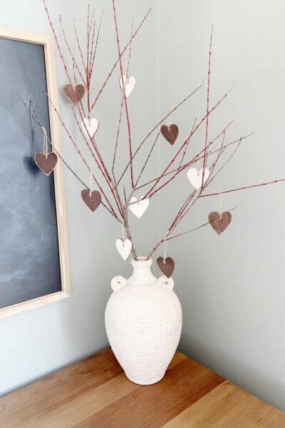 Painted heart ornaments hanging from a branch in a white vase.